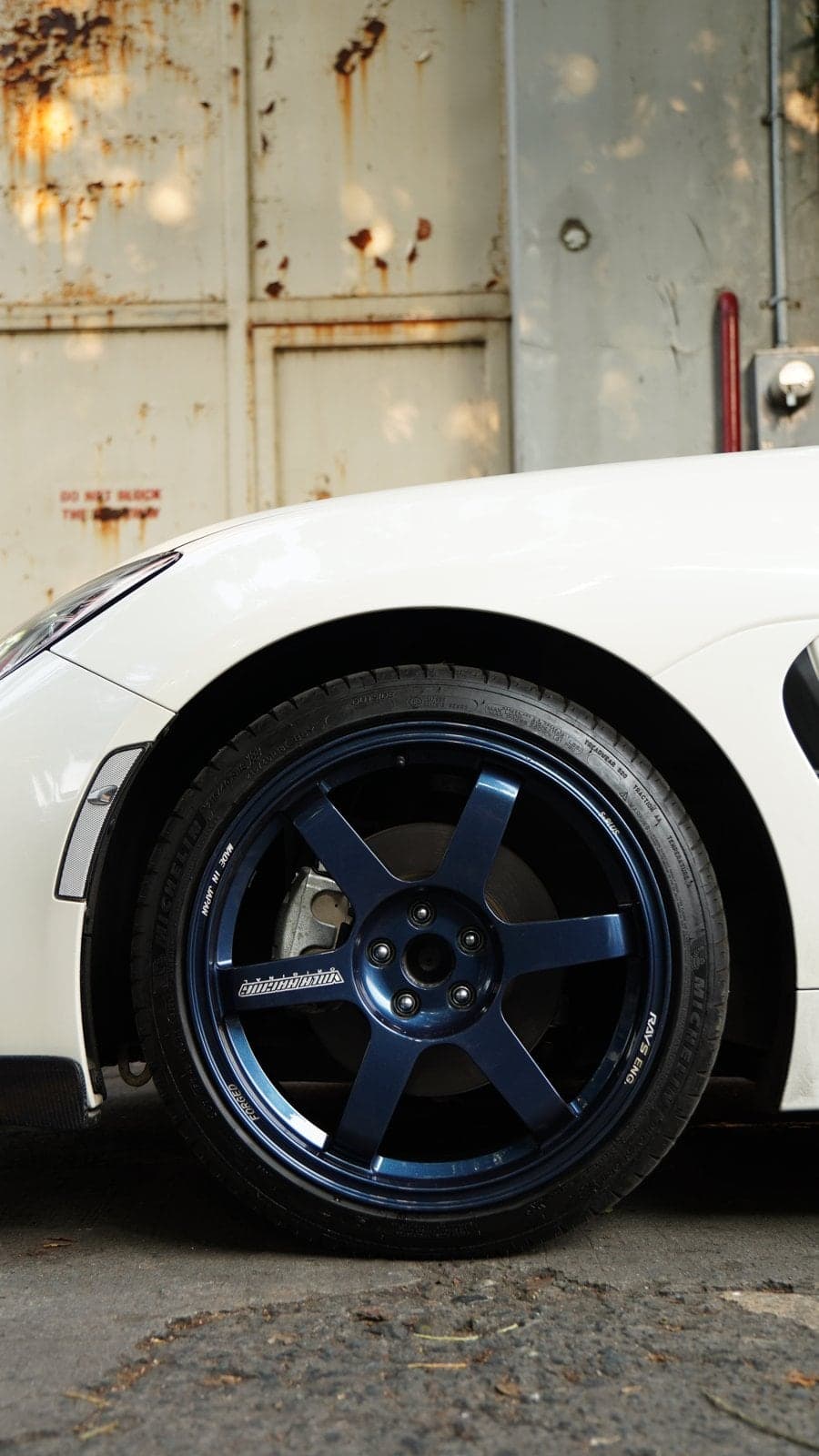 Blue six-spoke forged wheel on a white Subaru BRZ in front of a weathered metal door